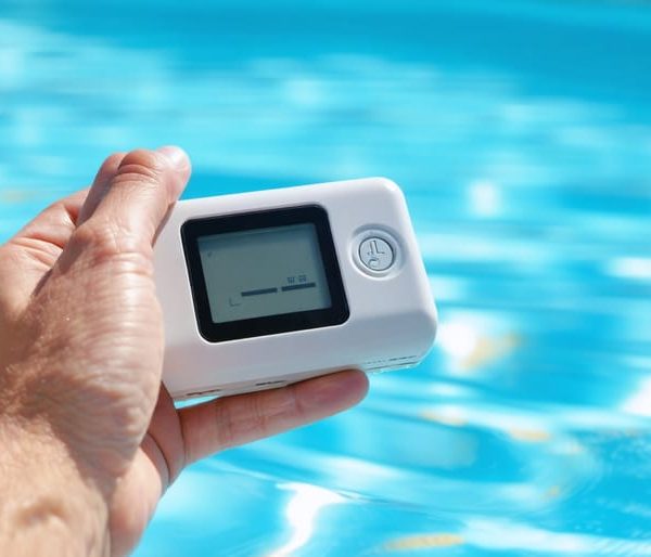 "Modern electronic pool water tester held by hand above a clear swimming pool, displaying digital readings."