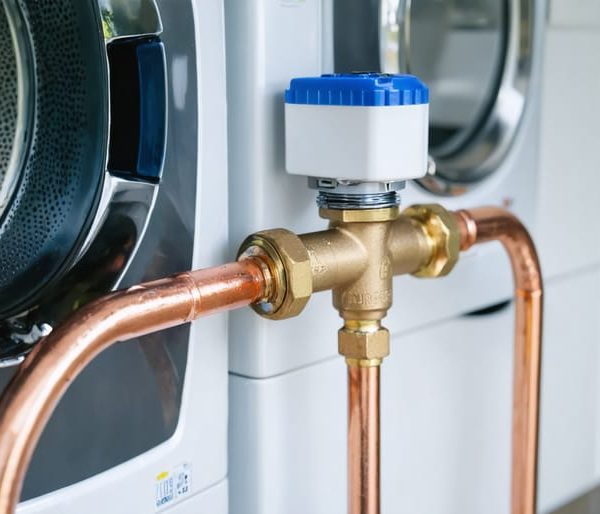 Close-up of a smart automatic water shutoff valve on a copper main supply line in a modern utility room, with washer and shelves softly blurred in the background.