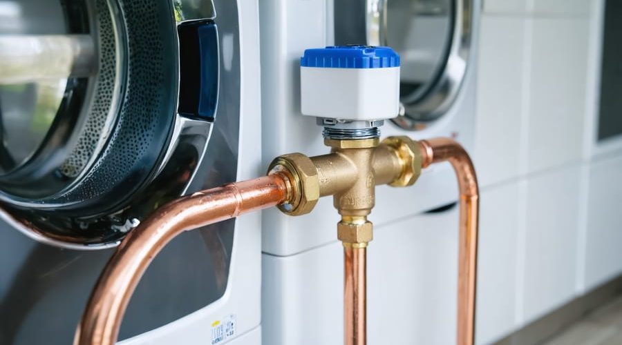 Close-up of a smart automatic water shutoff valve on a copper main supply line in a modern utility room, with washer and shelves softly blurred in the background.