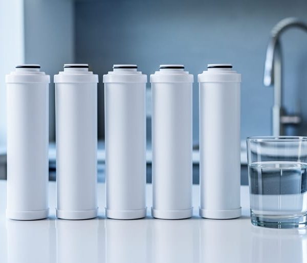 Five unbranded reverse osmosis filter cartridges in a neat row on a white countertop next to a clear glass of water, with a blurred stainless faucet and kitchen backsplash in the background under soft daylight