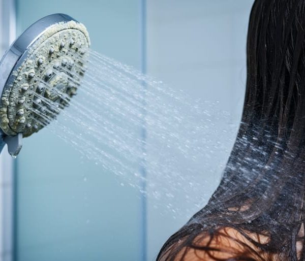 Close-up eye-level view of a limescale-coated chrome showerhead spraying onto wet, dull hair strands, with cool lighting and blurred white bathroom tiles in the background.